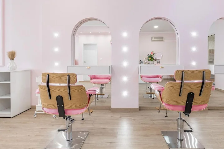 Cabeleireiro luminoso em rosa suave, com cadeiras de cabeleireiro em madeira e tecido rosa em frente a duas alcovas em arco iluminadas por focos, mobiliário branco e pavimento com efeito madeira clara.