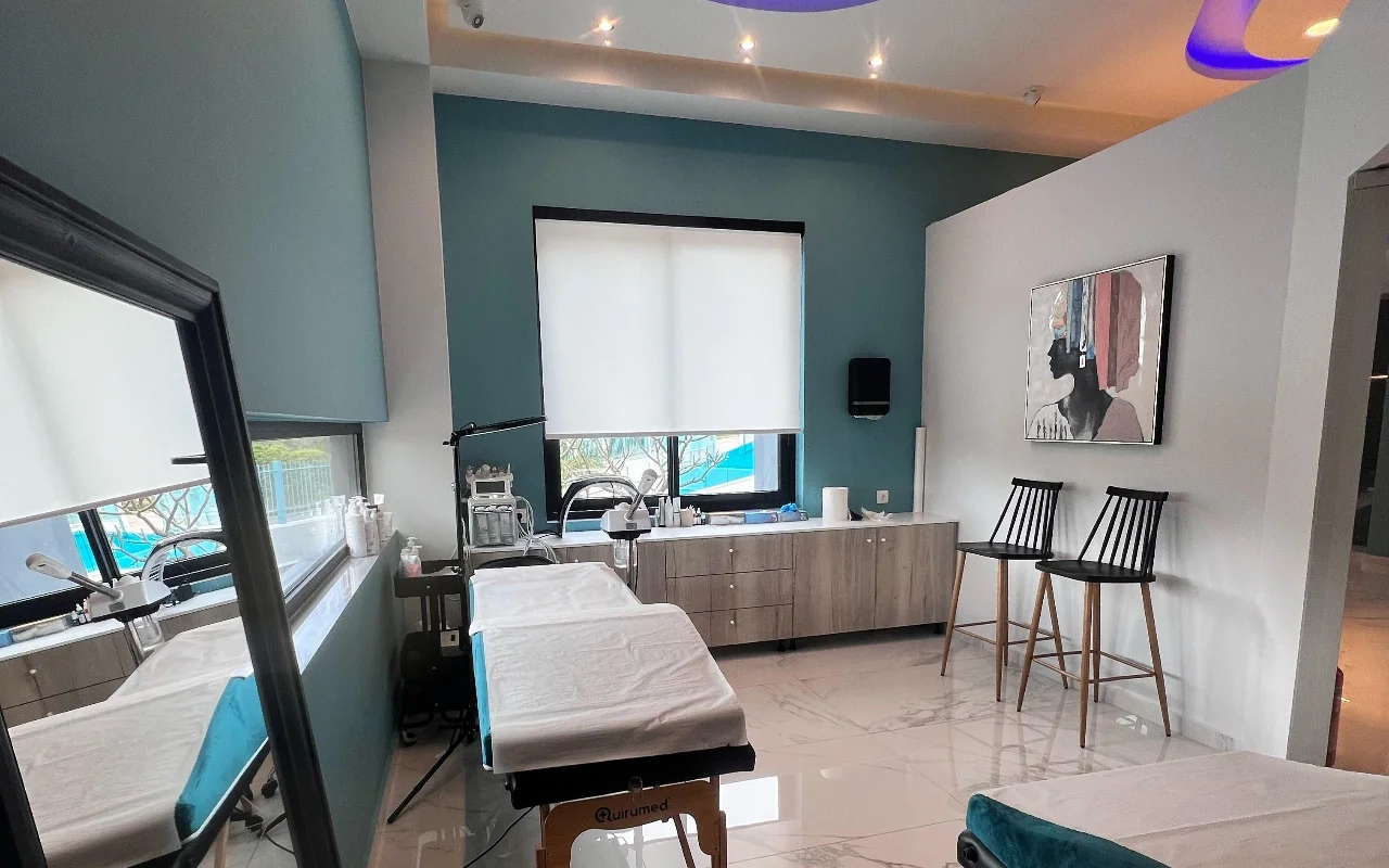 Uma sala de tratamento de rosto com paredes verde-petróleo, uma marquesa branca, aparelhos de skincare, um espelho grande, dois bancos pretos de ripas e um retrato enquadrado, iluminada por uma janela com estore branco de rolo.