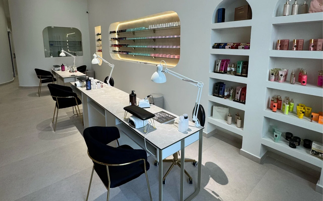 Um estúdio de unhas branco com mesas de manicure com tampo de mármore e candeeiros articulados brancos, cadeiras de veludo preto sobre pés dourados, e nichos de parede em arco a expor produtos de skincare e um expositor retroiluminado de vernizes.
