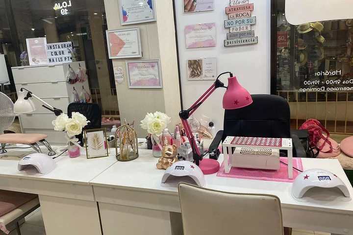 Estação de manicure de um salão de unhas com secretárias brancas, uma lâmpada rosa vibrante e lâmpadas LED, rosas brancas e certificados emoldurados na parede, com o nome Color Nails PT visível na montra ao lado do horário de abertura.