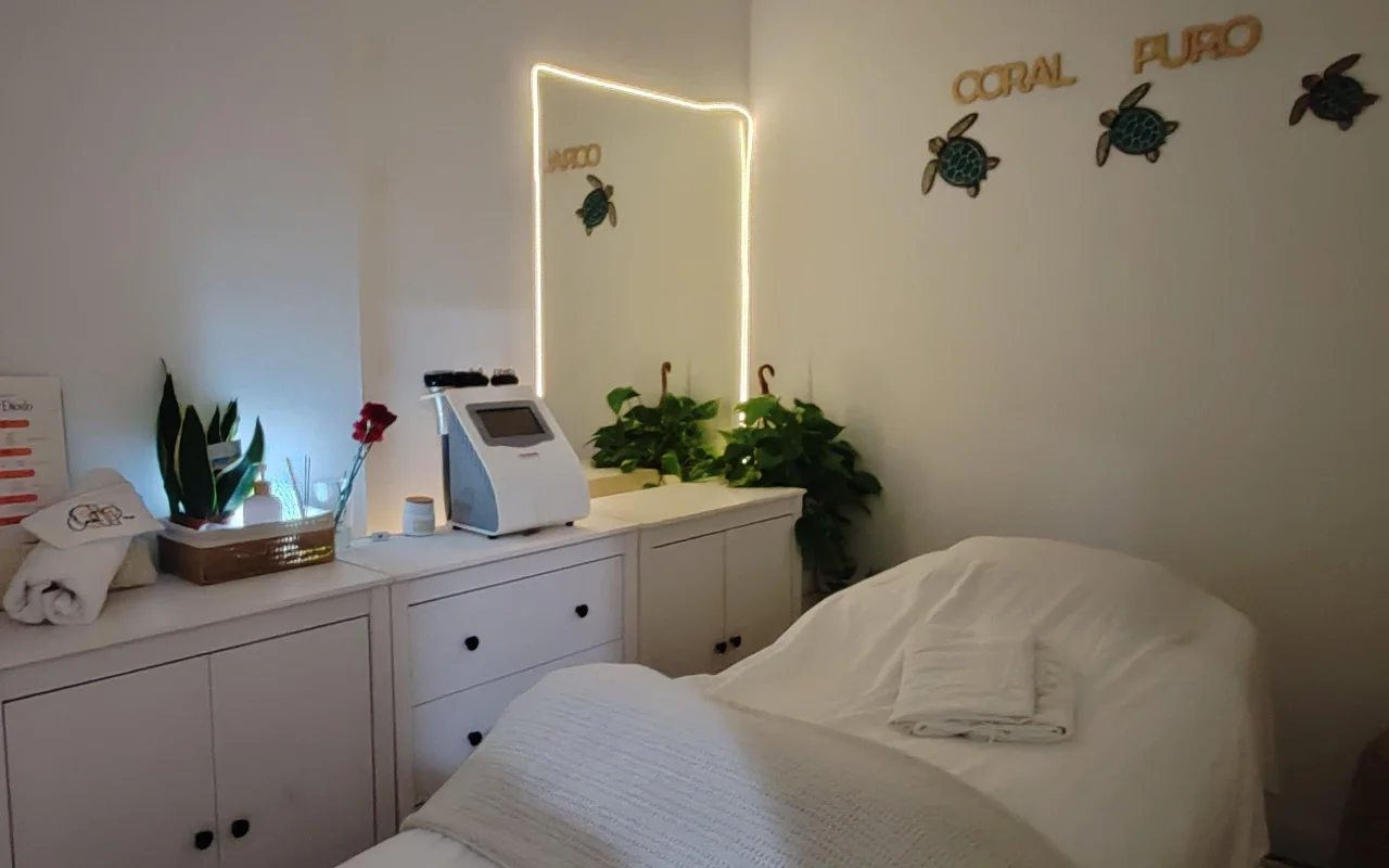 Uma pequena e calma sala de tratamentos com armários brancos e uma cama coberta com toalhas brancas. Um espelho iluminado e um aparelho de tratamento estão sobre o balcão entre plantas verdes, sob o letreiro dourado «Coral Puro» e arte de parede decorativa em forma de tartaruga.