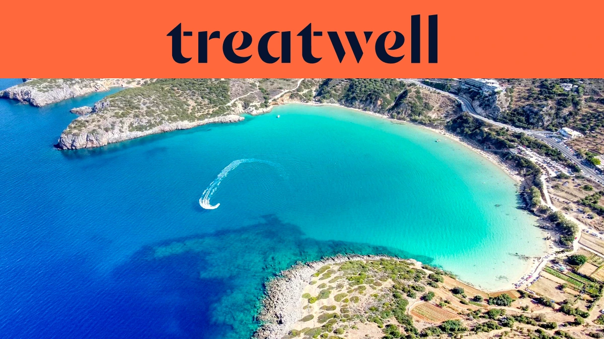 Vista aérea de uma baía cretense turquesa, com uma mota de água a traçar uma curva branca pela água limpa, uma meia-lua de praia de areia e colinas secas ao redor da margem, sob um banner cor de coral com a palavra treatwell.