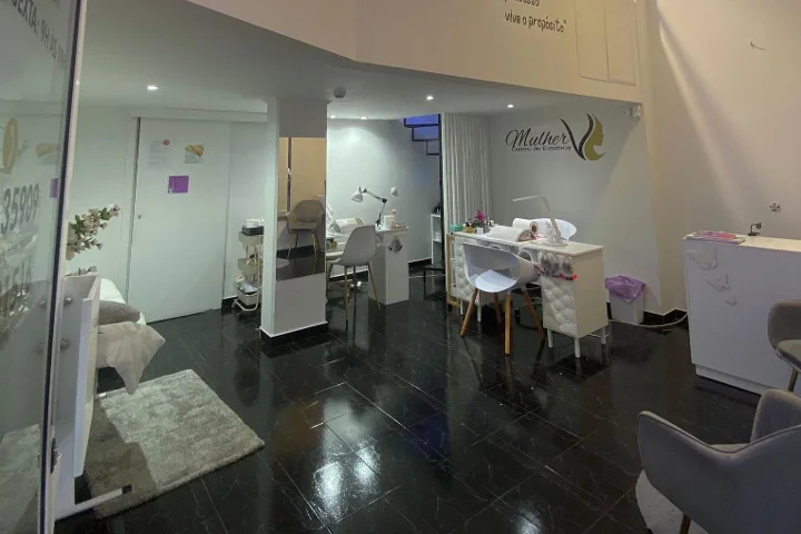 Interior de um centro de beleza: chão de ladrilho escuro e polido, estação de manicure branca e áreas de tratamento com luz suave, o logótipo Mulher na parede do fundo e uma marquesa de tratamento facial visível atrás de uma divisória de vidro.