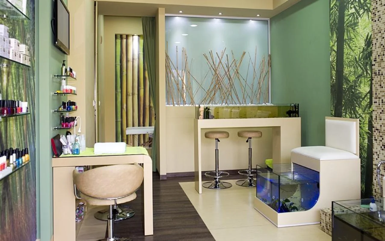 Um salão compacto com decoração de parede em bambu e verde-floresta, prateleiras de parede alinhadas de vernizes, uma mesa e cadeira de manicure creme, dois bancos de bar, e um aquário de vidro iluminado a azul para a pedicure fish spa.