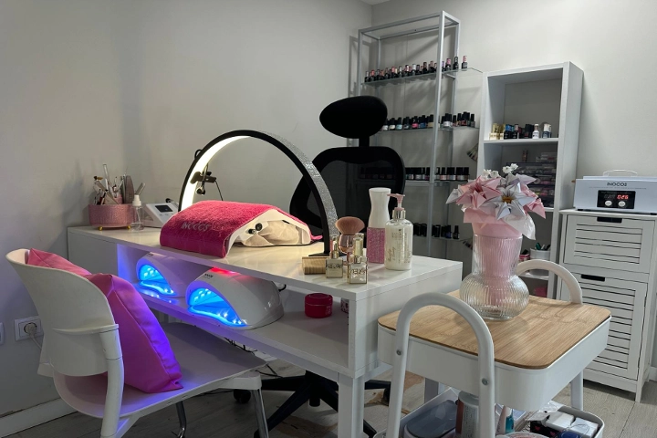 Posto único de manicure com uma secretária branca, uma ring light e duas lâmpadas LED azul-brilhantes, uma cadeira preta e prateleiras carregadas de vernizes coloridos, alegrado por um jarro rosa com flores.