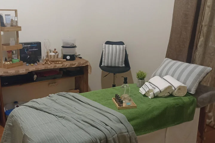 Gabinete de massagem acolhedor com uma marquesa coberta por uma toalha verde e toalhas brancas enroladas, uma bandeja de óleos essenciais, ferramentas de massagem em madeira sobre uma mesa rústica e um banco junto a um canto separado por uma cortina.