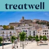 Vista ensolarada sobre Ibiza-cidade, com edifícios caiados de branco a subir até à catedral e às antigas muralhas fortificadas de Dalt Vila na colina. Um passeio portuário ladeado por árvores atravessa o primeiro plano sob um céu azul limpo.