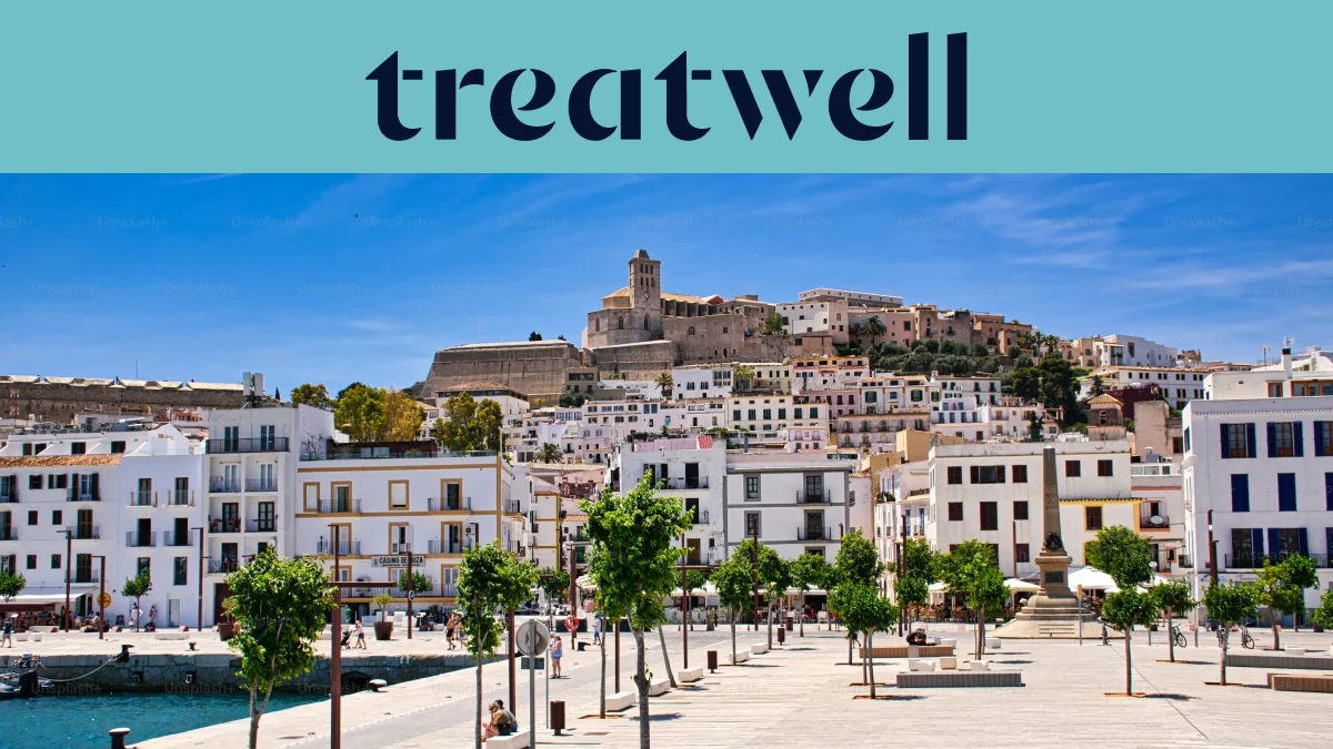 Vista ensolarada sobre Ibiza-cidade, com edifícios caiados de branco a subir até à catedral e às antigas muralhas fortificadas de Dalt Vila na colina. Um passeio portuário ladeado por árvores atravessa o primeiro plano sob um céu azul limpo.