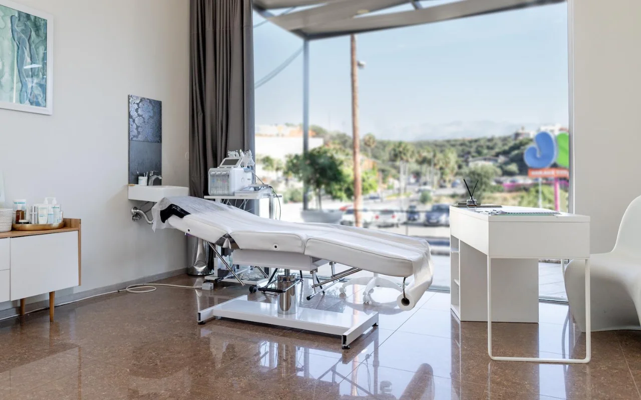 Uma sala de tratamento branca e imaculada com uma marquesa de estética reclinável, equipamento de skincare num carrinho, obras enquadradas e uma janela grande com vista para árvores, uma estrada e colinas ao longe.