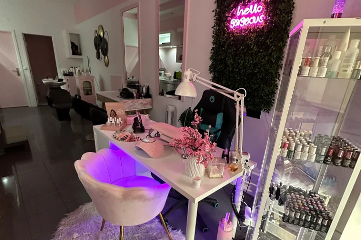 Posto de beleza iluminado a rosa, com um letreiro néon 'hello gorgeous' numa parede vegetal, uma cadeira creme em forma de concha, uma secretária branca com pincéis e flores de cerejeira, e uma vitrina cheia de vernizes coloridos.