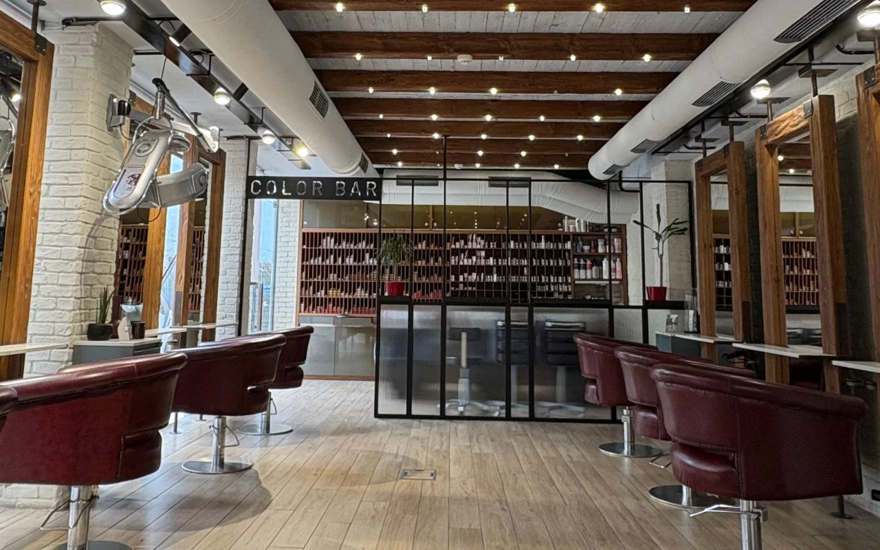Um salão de cabeleireiro com vigas de madeira à vista e tijolo caiado de branco, cadeiras de styling de couro bordô, e uma sala de cor com paredes envidraçadas revestida de prateleiras de produtos atrás de um letreiro 'Color Bar'.