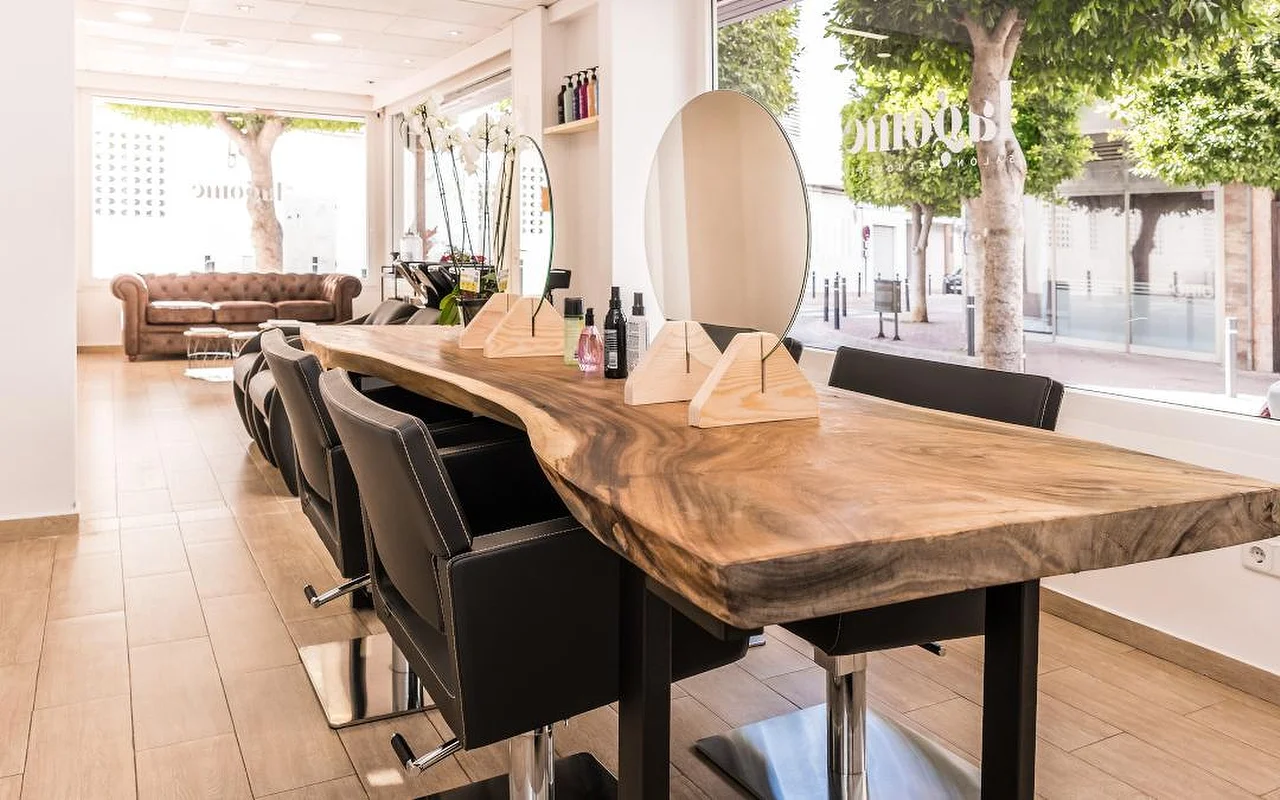 Um salão de cabelo luminoso construído em torno de um longo balcão de madeira com bordo natural, alinhado com cadeiras de styling pretas. Um espelho redondo e suportes de madeira para produtos ficam sobre o balcão, um sofá Chesterfield em pele castanha aguarda ao fundo, e altas janelas dão para uma rua arborizada.