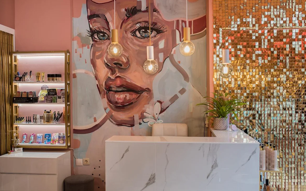 Um salão de paredes cor-de-rosa com um grande mural pintado do rosto de uma mulher, uma parede de lantejoulas douradas brilhantes, um balcão de receção em mármore branco e candeeiros de teto com lâmpadas à vista.