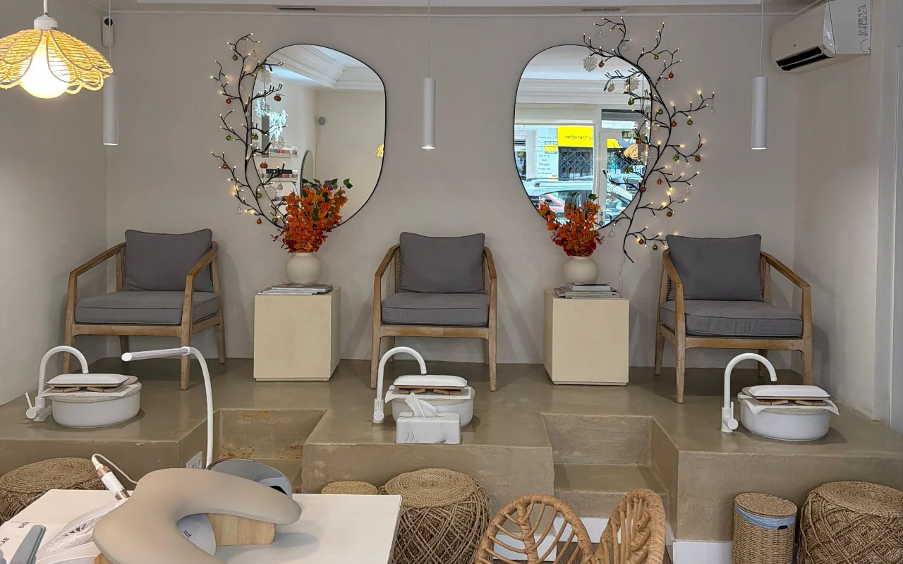 Um salão de unhas luminoso e moderno com duas cadeiras de pedicura numa plataforma de betão elevada, cada uma com um pé-de-banho branco e uma poltrona cinza almofadada. Dois espelhos de formas orgânicas, decorados com ramos iluminados e flores cor de laranja, pendem por cima, com um posto de manicura em primeiro plano.