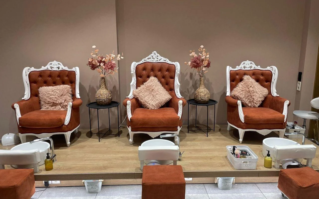 Três cadeirões ornamentados com molduras brancas em veludo cor de laranja queimada, elevados numa plataforma de madeira sobre bacias de pedicure brancas, com flores secas e almofadas rosa suave entre os assentos.