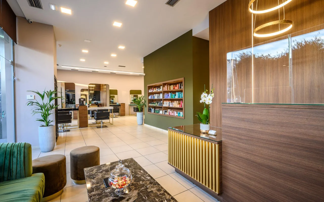 Um salão de cabeleireiro sofisticado com painéis de madeira quente, uma parede decorativa verde-azeitona com prateleiras de produtos, postos de styling com espelhos grandes, um balcão de receção com ripas douradas e assentos de veludo verde sob candeeiros suspensos dourados circulares.
