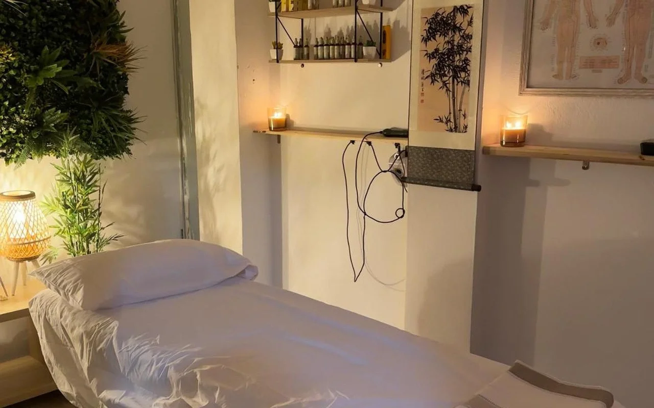 Uma sala de tratamentos suavemente iluminada com uma cama de massagem feita em linho branco. Velas acesas descansam em prateleiras de parede em madeira ao lado de um rolo de tinta em bambu e uma tabela de acupuntura emoldurada, com uma planta verdejante e um candeeiro entrançado luminoso no canto.