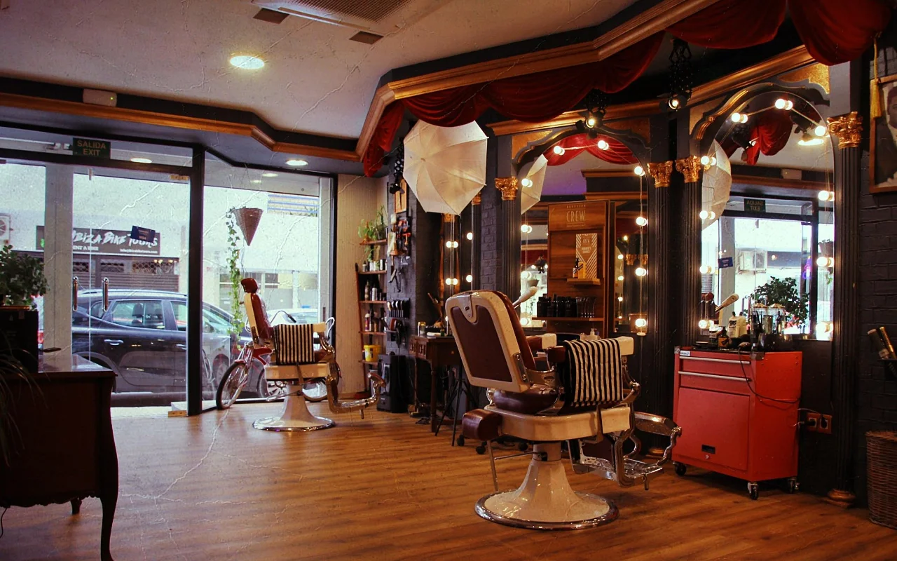 Uma barbearia de estilo vintage com clássicas cadeiras reclináveis em pele sobre bases cromadas e toalhas às riscas. Espelhos dourados enquadrados por luzes redondas em bola e drapeados de veludo vermelho revestem a parede, um carrinho de ferramentas vermelho fica a um lado, e janelas viradas para a rua iluminam o soalho de madeira.