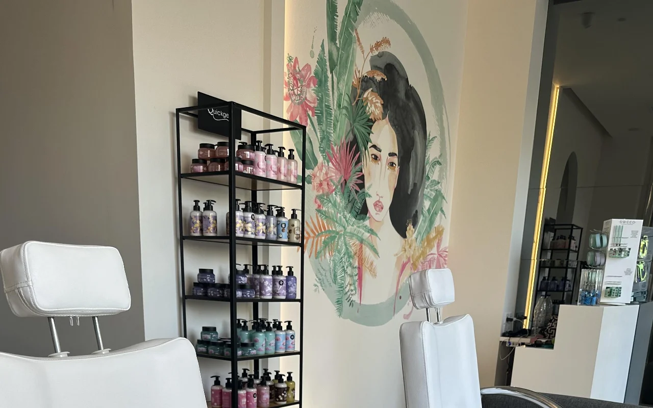 Um canto de estúdio com duas cadeiras de tratamento brancas reclináveis, uma prateleira preta de produtos, um espelho retroiluminado, e um mural floral do rosto de uma mulher enquadrado por folhagem tropical.