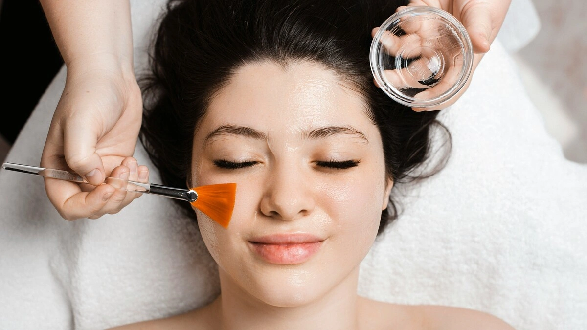 Vista de cima de uma esteticista a aplicar uma solução de peeling químico no rosto de uma cliente com um pincel largo e plano