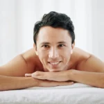 Um homem está deitado de barriga para baixo na marquesa de um centro de estética, com o queixo apoiado nas mãos, e sorri