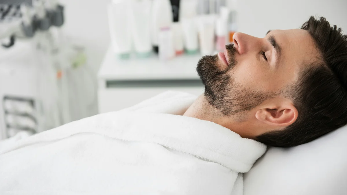 Um homem está deitado na marquesa com um roupão branco, a cabeça inclinada para trás e os olhos fechados