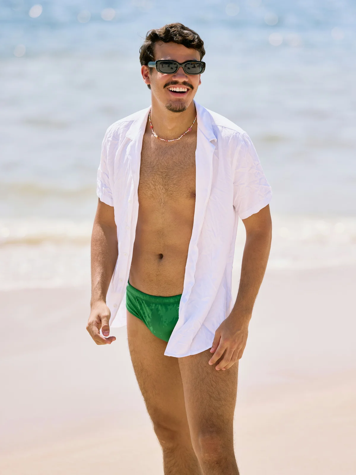 Um homem sorridente está de pé numa praia num dia solarengo; veste uns calções de banho verdes, uma camisa branca aberta e óculos de sol pretos