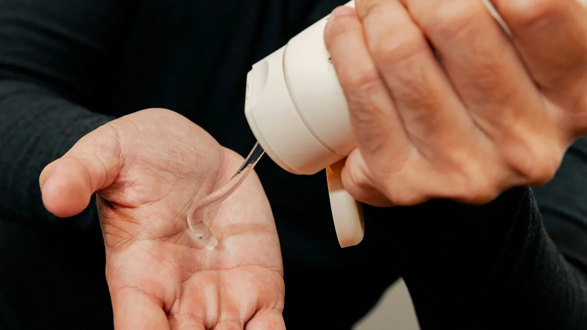 Grande plano das mãos de um homem que, com uma mão, espreme um tubo branco de gel hidratante para a outra mão
