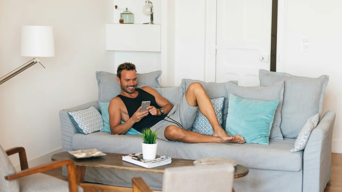 Um homem relaxa com os pés para cima no sofá e sorri enquanto olha para o telemóvel; veste uma camisola de alças preta e uns calções cinzentos largos e macios