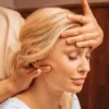 Grande plano de uma mulher com os olhos fechados enquanto uma profissional, durante uma massagem indiana à cabeça, lhe apoia uma mão na testa e com a outra lhe massaja o pescoço