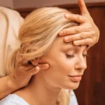 Grande plano de uma mulher com os olhos fechados enquanto uma profissional, durante uma massagem indiana à cabeça, lhe apoia uma mão na testa e com a outra lhe massaja o pescoço