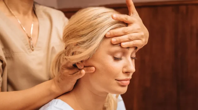 Grande plano de uma mulher com os olhos fechados enquanto uma profissional, durante uma massagem indiana à cabeça, lhe apoia uma mão na testa e com a outra lhe massaja o pescoço