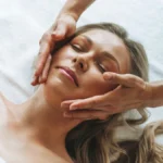 Mulher relaxada durante uma massagem suave de drenagem linfática ao rosto, com as mãos da profissional a deslizar levemente sobre a face
