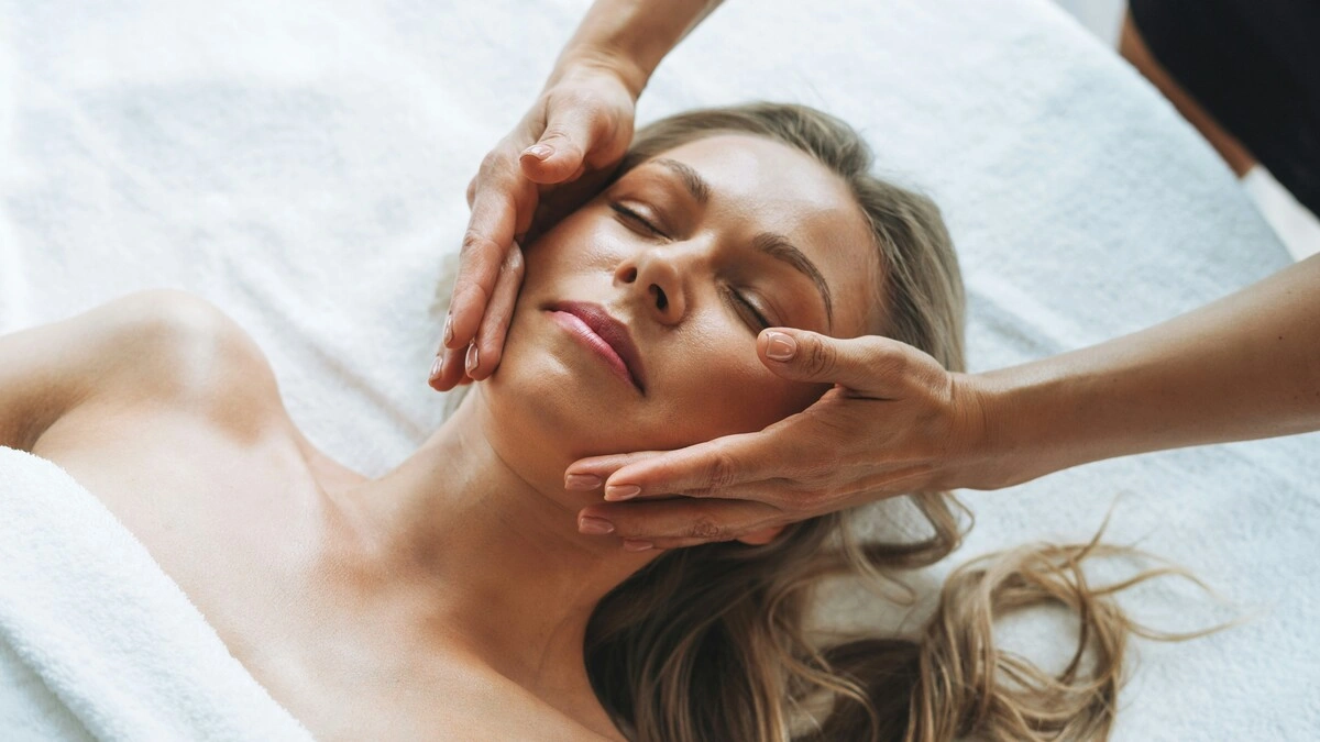 Mulher relaxada durante uma massagem suave de drenagem linfática ao rosto, com as mãos da profissional a deslizar levemente sobre a face