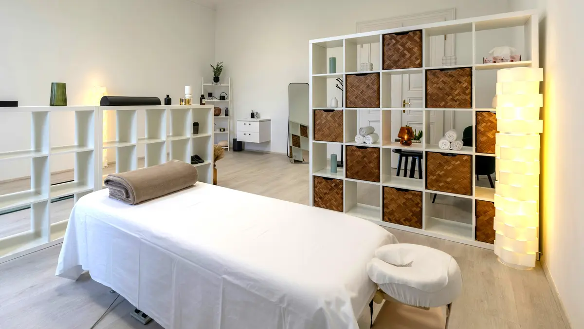 Espaço de tratamentos limpo e arrumado num salão, com a marquesa preparada com lençóis e uma manta enrolada, pronta para uma massagem